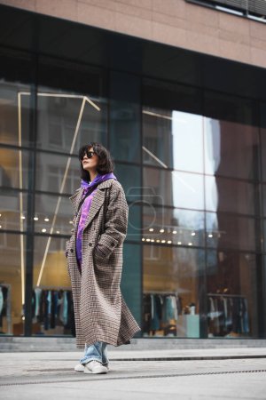 Photo for Elegant casual outfit. Beautiful young woman in trendy sunglasses, wearing coat with colorful hoodie, posing against modern city building. Concept of street style fashion, beauty, modern trends - Royalty Free Image