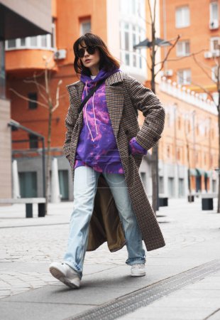 Téléchargez les photos : Défilé de mode urbain. Jeune femme en pose dynamique portant un manteau, un jean et un sweat à capuche lumineux marchant le long de la rue vide. Tenue décontractée. Concept de mode street style, beauté, tendances modernes - en image libre de droit