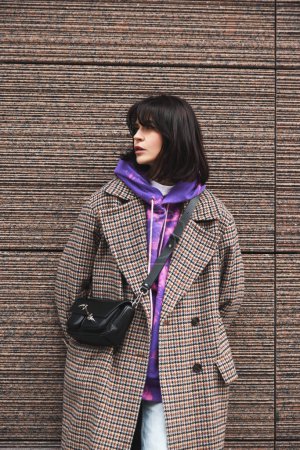 Téléchargez les photos : Portrait de jeune femme brune portant un manteau décontracté élégant et un sweat à capuche lumineux, posant contre l'architecture urbaine. Confortable, chaleureux et élégant. Concept de mode street style, beauté, tendances modernes - en image libre de droit
