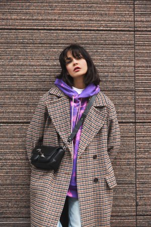 Téléchargez les photos : Portrait de jeune femme brune portant un manteau décontracté élégant et un sweat à capuche lumineux, posant contre l'architecture urbaine. Confortable, chaleureux et élégant. Concept de mode street style, beauté, tendances modernes - en image libre de droit