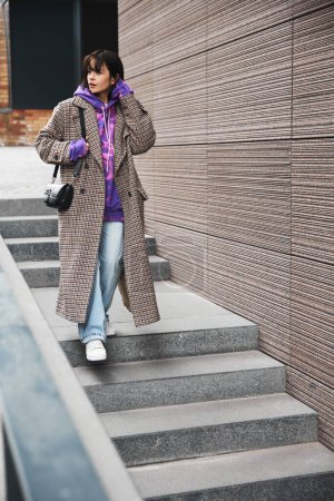 Téléchargez les photos : Elle arrive. Jeune femme élégante descendant les escaliers, portant timide à la mode, sweat à capuche violet coloré et jean décontracté. Style urbain. Concept de mode street style, beauté, tendances modernes - en image libre de droit