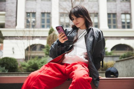 Photo for Young woman in stylish gray jacket and red pants sitting on bench with mobile phone against urban background. Concept of street style fashion, beauty, modern trends - Royalty Free Image