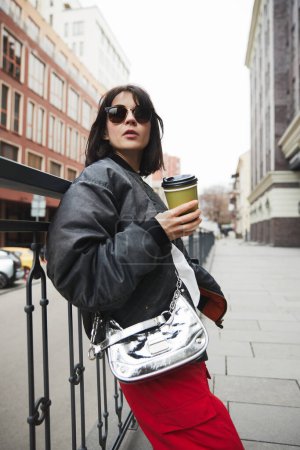 Photo for Young woman in stylish gray jacket, metal colored bag and red pants standing with coffee cup, walking around city. Concept of street style fashion, beauty, modern trends - Royalty Free Image
