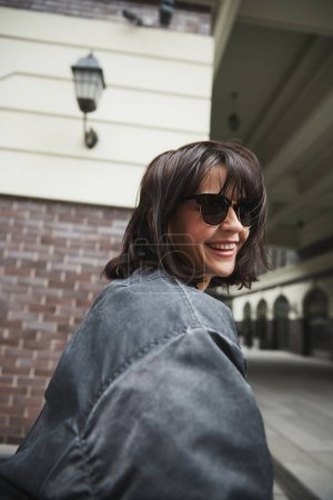Téléchargez les photos : Portrait de belle jeune femme brune en lunettes de soleil et veste tendance décontractée grise se promenant en ville, posant. Concept de mode street style, beauté, tendances modernes - en image libre de droit