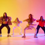 Four artistic young women in sport style casual clothes in motion, dancing freestyle dance against gradient background in neon. Concept of youth, street dance, contemporary dance, modern, dynamics
