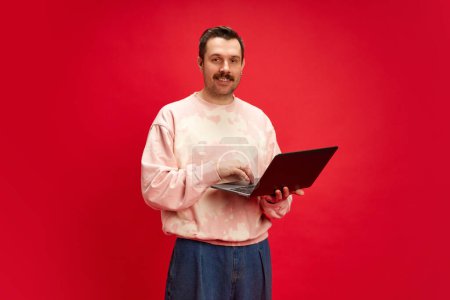 Foto de Hombre con sudadera de pie con portátil aislado en el fondo rojo del estudio. Software de trabajo remoto que demuestra una comunicación fácil. Concepto de emociones humanas, moda casual, estilo de vida, negocios - Imagen libre de derechos