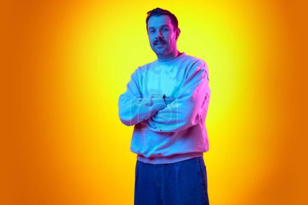 Photo for Man in his 30s wearing sweatshirt and jeans, standing with arms crossed on gradient yellow orange background in neon light. Relaxed. Concept of human emotions, casual fashion, lifestyle - Royalty Free Image