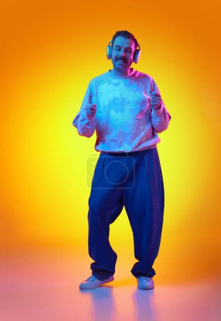Foto de Hombre positivo, enérgico y sonriente en sudadera rosa y jeans escuchando música en auriculares y bailando sobre un fondo naranja amarillento degradado en neón. Concepto de emociones humanas, moda casual - Imagen libre de derechos
