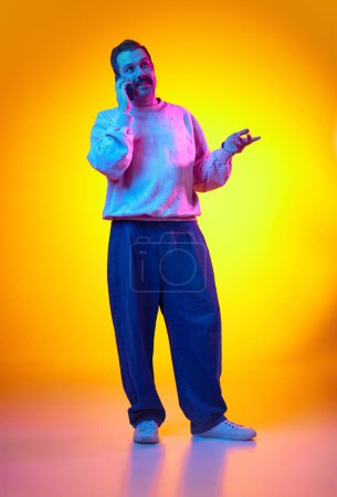 Photo for Young smiling man in sweatshirt and jeans talking on mobile phone and gesturing on gradient yellow orange background in neon light. Concept of human emotions, casual fashion, lifestyle - Royalty Free Image