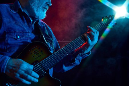 Photo for Senior bearded man playing guitar against dark background with dusk element. Concept, performance. Concept of music, instruments, concert, sound, equipment, festival - Royalty Free Image