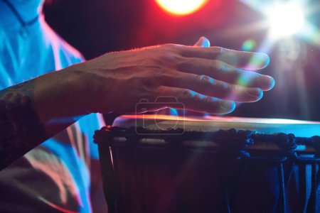 Foto de Primer plano de manos masculinas, músico tocando tambores de bongo sobre fondo oscuro con luces de escenario. Cultura africana evento en vivo. Concepto de música, instrumentos, concierto, sonido, equipo, festival - Imagen libre de derechos