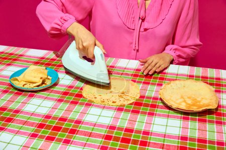 Foto de Mujer en blusa rosa planchando panqueque sobre mantel a cuadros. Meme con formas inesperadas de preparar la comida a través del humor. Concepto de arte pop, creatividad, rareza, comida, humor, fotografía colorida - Imagen libre de derechos