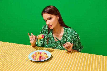 Téléchargez les photos : Jeune femme mangeant des spaghettis sur des bigoudis sur fond vert. Présentation inhabituelle de la nourriture. Menu moderne. Concept de cuisine pop art photographie, créativité, style décalé - en image libre de droit
