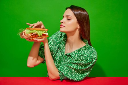 Foto de Mujer joven comiendo sándwich extravagante con muñeca sobre fondo rojo. Lechuga, tomate y ketchup para un sabor adicional. Concepto de comida pop arte fotografía, creatividad, estilo peculiar - Imagen libre de derechos