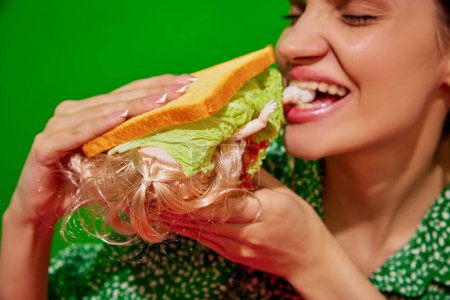 Foto de Mujer joven comiendo sándwich extravagante con muñeca sobre fondo rojo. Extrañeza y surrealismo. Concepto de comida pop arte fotografía, creatividad, estilo peculiar - Imagen libre de derechos