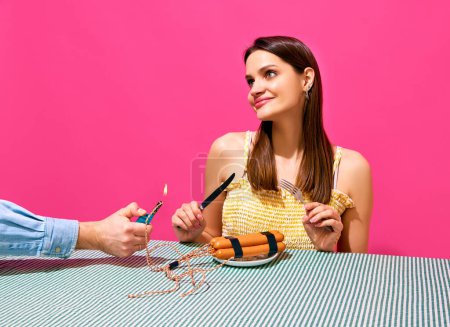 Foto de Mujer joven sonriente sentada en la mesa con jugador y salchichas imitando artefacto explosivo sobre fondo rosa. Surrealismo. ¡Bang! Concepto de comida pop arte fotografía, creatividad, estilo peculiar - Imagen libre de derechos