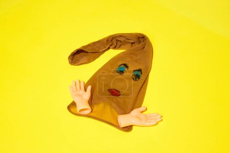 Pantyhose with face drawn on it, toy hands extending from sides, on against bright yellow background. DIY fashion brand. Concept of food pop art photography, creativity, quirky style
