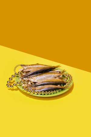 Téléchargez les photos : Poisson fumé sur une assiette avec des colliers de perles, sur un fond jaune en diagonale, arrangement élégant. Concept de cuisine pop art photographie, créativité, style décalé - en image libre de droit
