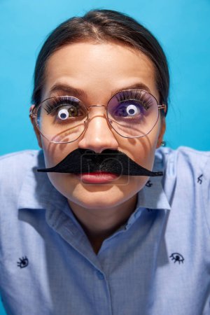 Foto de Mujer joven con camisa, con bigotes falsos, gafas con ojos en ella y haciendo expresiones divertidas contra el fondo azul. Concepto de comida pop arte fotografía, creatividad, estilo peculiar - Imagen libre de derechos