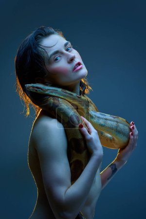 Foto de Retrato de una mujer joven y elegante con mejillas rosas posando sin camisa con serpiente sobre su cuerpo sobre fondo de estudio azul oscuro en neón. Concepto de belleza femenina, tema animal, cuidado, elegancia, moda - Imagen libre de derechos
