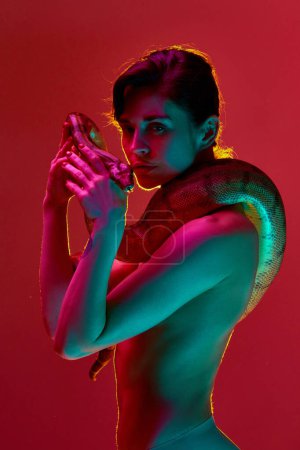 Photo for Fantasy and dreams. Beautiful young shirtless woman with slim body posing with snake against red studio background in neon light. Concept of female beauty, animal theme, care, elegance, fashion - Royalty Free Image