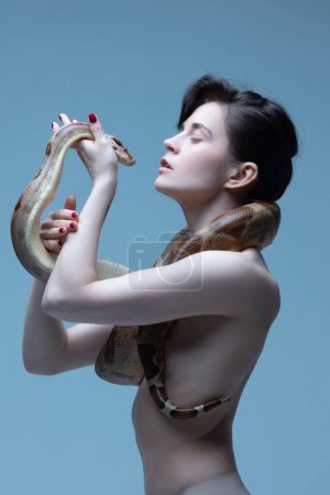 Téléchargez les photos : Proche de la nature. Belle jeune femme brune au corps mince posant avec serpent sur fond de studio bleu. Concept de beauté féminine, thème animal, soins, élégance, mode - en image libre de droit