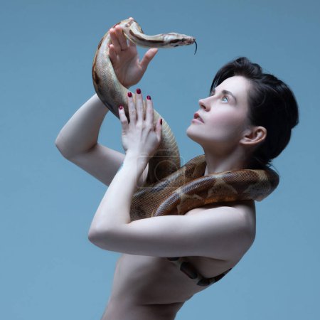 Foto de Hermosa joven morena con cuerpo delgado posando con serpiente sobre fondo de estudio azul. Interacción humana con animales. Concepto de belleza femenina, tema animal, cuidado, elegancia, moda - Imagen libre de derechos