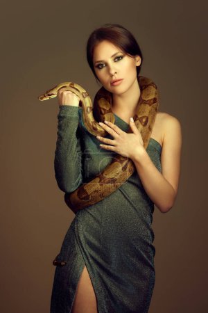 Téléchargez les photos : Jeune femme en robe verte pailletée avec maquillage, posant avec serpent sur fond de studio brun. Soirée porter des campagnes de mode. Concept de beauté féminine, thème animal, soins, élégance, mode - en image libre de droit