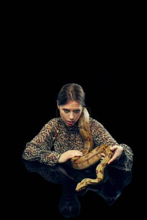 Foto de Retrato de mujer joven con maquillaje, en blusa animal estampada posando con serpiente sobre fondo de estudio negro. Concepto de belleza femenina, tema animal, cuidado, elegancia, moda - Imagen libre de derechos