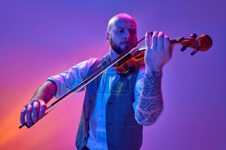 Photo for Bald man in classical clothes, with tattoo, musician playing violin against gradient purple background in neon light. Concept of music, talent show, performance, concert, festival, instruments - Royalty Free Image