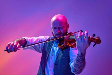 Photo for Bald man in classical clothes, with tattoo, musician playing violin against gradient purple background in neon light. Concept of music, talent show, performance, concert, festival, instruments - Royalty Free Image