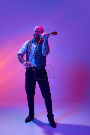 Photo for Full-length of emotional bearded bald man, in classical suit playing violin against gradient purple background in neon light. Concept of music, talent show, performance, concert, festival, instruments - Royalty Free Image