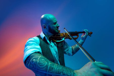 Photo for Deep feelings of melody. Young bald man playing violin against blue background in neon with mixed light. Concept of music, talent show, performance, concert, festival, instruments - Royalty Free Image