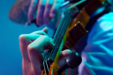 Cropped image of male hands plying violin. Man making performance against blue background in neon with mixed light. Concept of music, talent show, performance, concert, festival, instruments