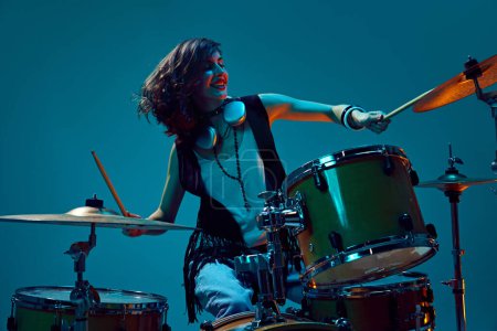 Photo for Beautiful, energetic young woman, artistic musician playing drums against cyan background in neon light. Concept of music, talent show, performance, concert, festival, instruments - Royalty Free Image
