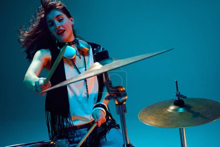 Photo for Beautiful young woman in stylish clothes, musician playing drums against cyan background in neon light. Rock and roll event. Concept of music, talent show, performance, concert, festival, instruments - Royalty Free Image