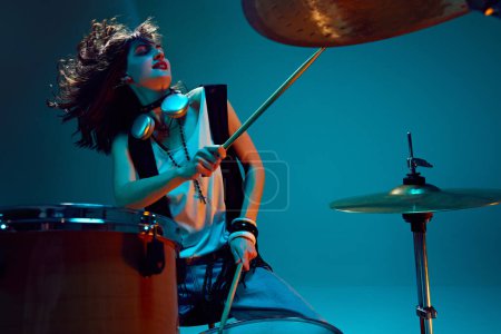 Photo for Young energetic beautiful woman in stylish clothes, musician playing drums against cyan background in neon light. Concept of music, talent show, performance, concert, festival, instruments - Royalty Free Image