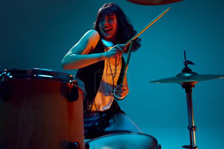Photo for Energy burst. Young emotional, active woman, musician in stylish clothes playing drums against cyan background in neon light. Concept of music, talent show, performance, concert, festival, instruments - Royalty Free Image