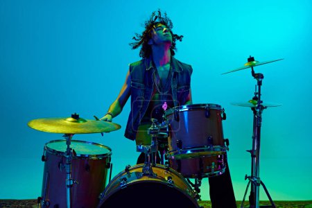 Photo for Stylish young man, musician playing drums, making solo performance against blue background in neon light. Concept of music, talent show, performance, concert, festival, instruments, youth culture - Royalty Free Image