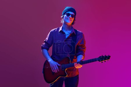Young man in casual clothes and sunglasses, playing guitar against pink background in neon light. Solo performer. Concept of music, talent show, performance, concert, festival, instruments