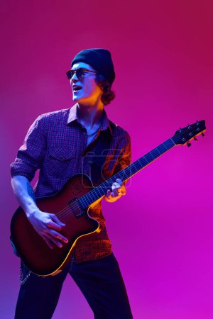 Talented, artistic young man in casual clothes and sunglasses, playing guitar against pink background in neon light. Concept of music, talent show, performance, concert, festival, instruments