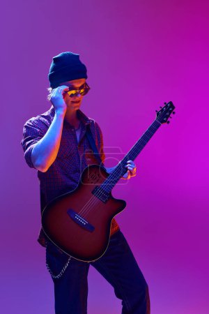 Foto de Guapo joven con estilo en gafas de sol, sombrero y ropa casual tocando la guitarra sobre fondo rosa en luz de neón. Concepto de música, show de talentos, performance, concierto, festival, instrumentos - Imagen libre de derechos