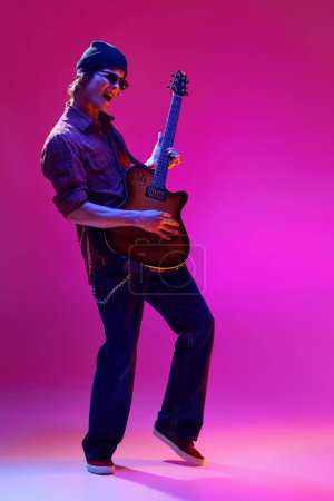 Handsome stylish young man in sunglasses, hat and casual clothes playing guitar against pink background in neon light. Concept of music, talent show, performance, concert, festival, instruments