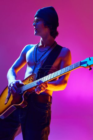 Handsome shirtless young man in sunglasses and hat playing electric guitar against pink background in neon light. Concept of music, talent show, performance, concert, festival, instruments