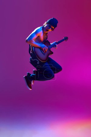 Energetic young shirtless man in sunglasses,rocket playing guitar against pink background in neon light. Concept of music, talent show, performance, concert, festival, instruments