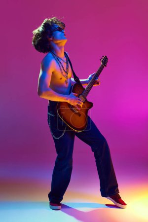 Rock and roll concert. Artistic and emotional shirtless young man playing guitar against pink background in neon light. Concept of music, talent show, performance, concert, festival, instruments
