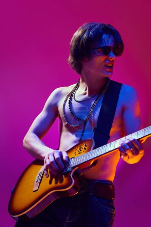 Handsome young shirtless man with accessories, sunglasses playing guitar against pink background in neon light. Concept of music, talent show, performance, concert, festival, instruments