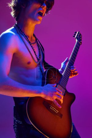 Talented and artistic young man, shirtless musician playing electric guitar against pink background in neon light. Concept of music, talent show, performance, concert, festival, instruments