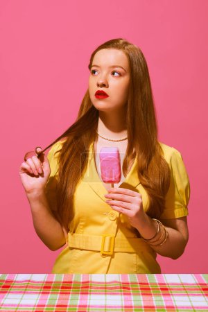 Photo for Redhead young woman with red lips, in yellow dress standing with ice cream and thoughtful face against pink background. Vintage-inspired dress collection. Concept of pop art photography, creativity - Royalty Free Image