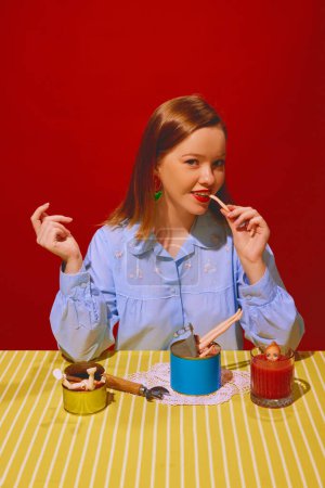 Photo for Young beautiful woman sitting at table and eating canned meat, drinking tomato juice against red background. Quirky food representation. Concept of pop art photography, creativity, food, weirdness - Royalty Free Image